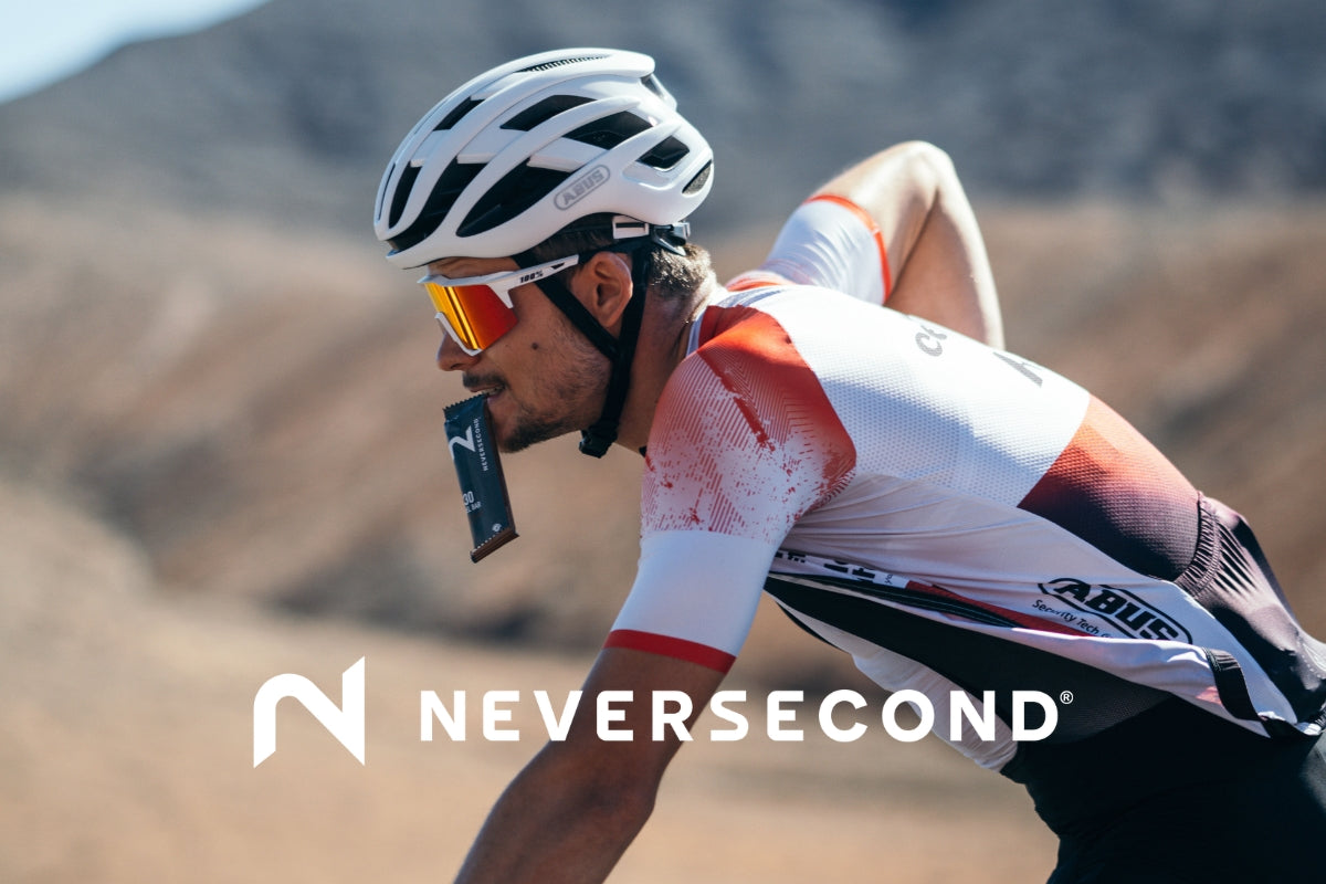 Triathlet Jan Stratmann fährt auf dem Rennrad durch eine karge Berglandschaft und hat ein Energiegel von der Marke Neversecond im Mund; im Vordergrund ist das Neversecond-Logo eingeblendet.