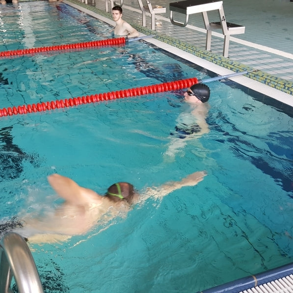 Zwei Schwimmer im Wasser, einer wartet am Beckenrad, der andere schwimmt gerade mit gestrecktem Arm heran